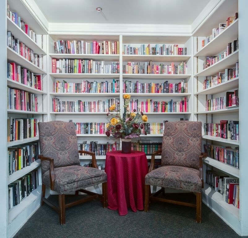 A cozy reading nook in a library with two armchairs, a small table, and shelves filled with books.