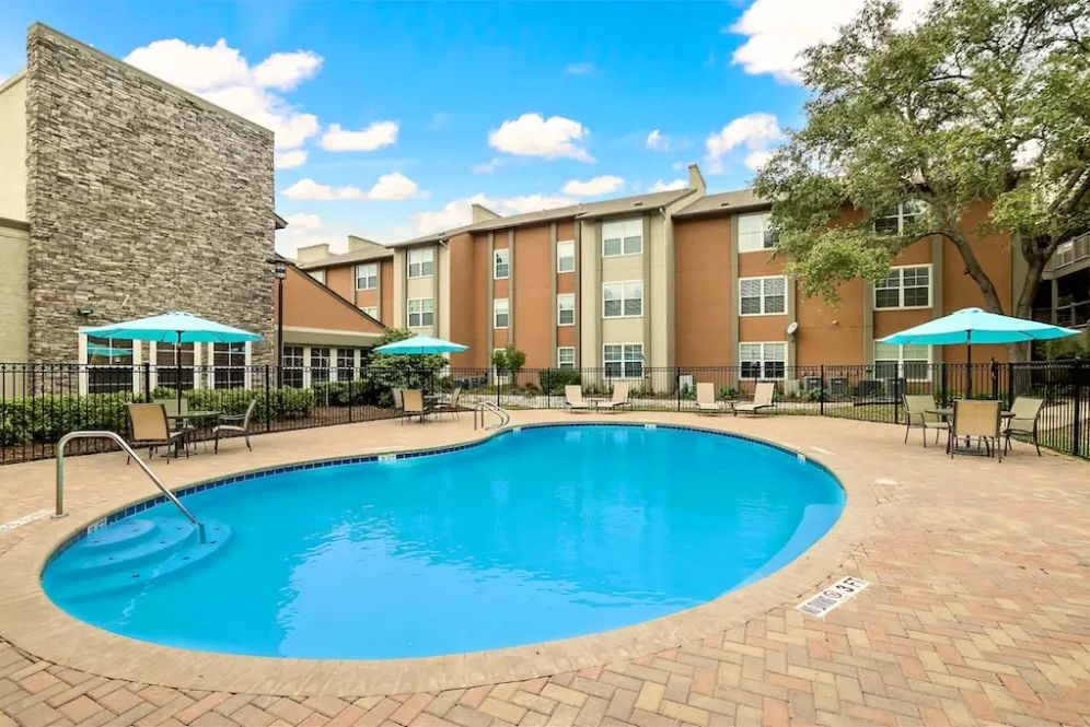 A tranquil swimming pool area surrounded by an apartment complex with patio furniture under a clear blue sky.