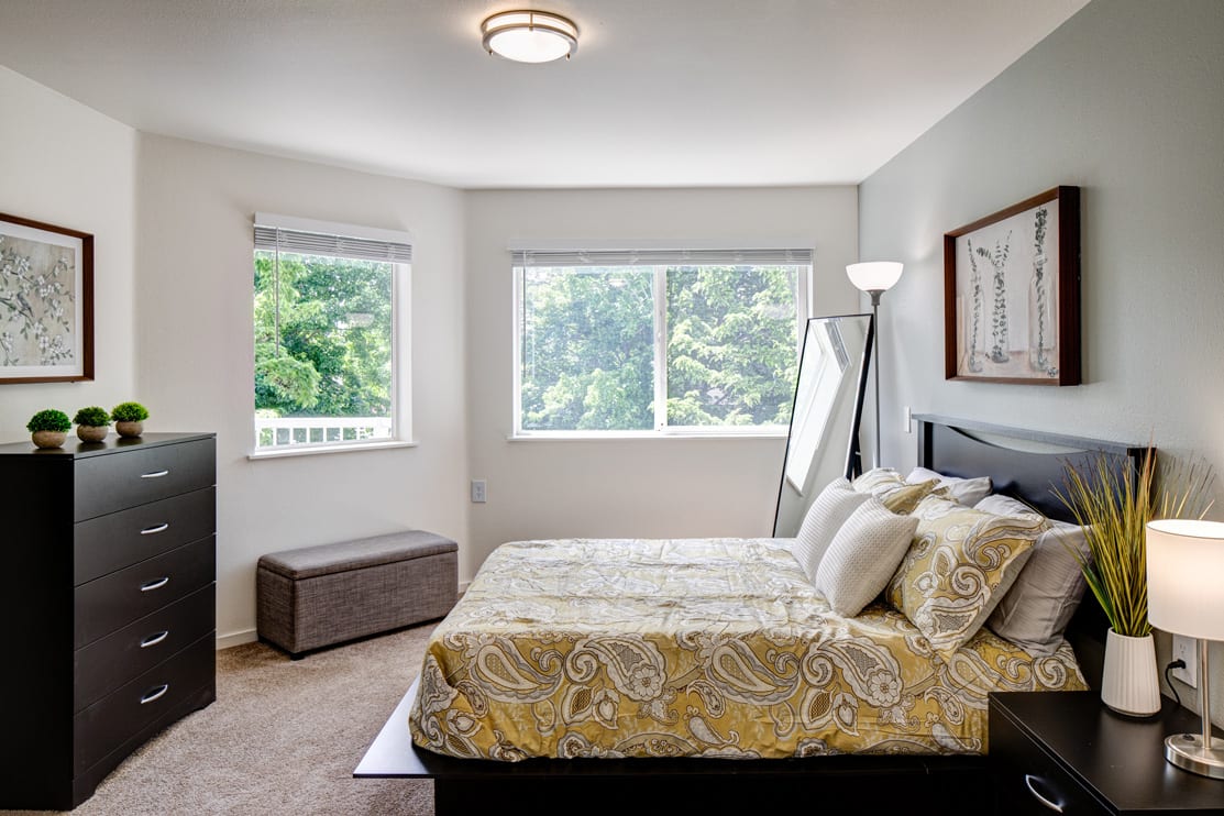 A bright and cozy bedroom with a large window, featuring a comfortable bed, a standing lamp, and a full-length mirror.