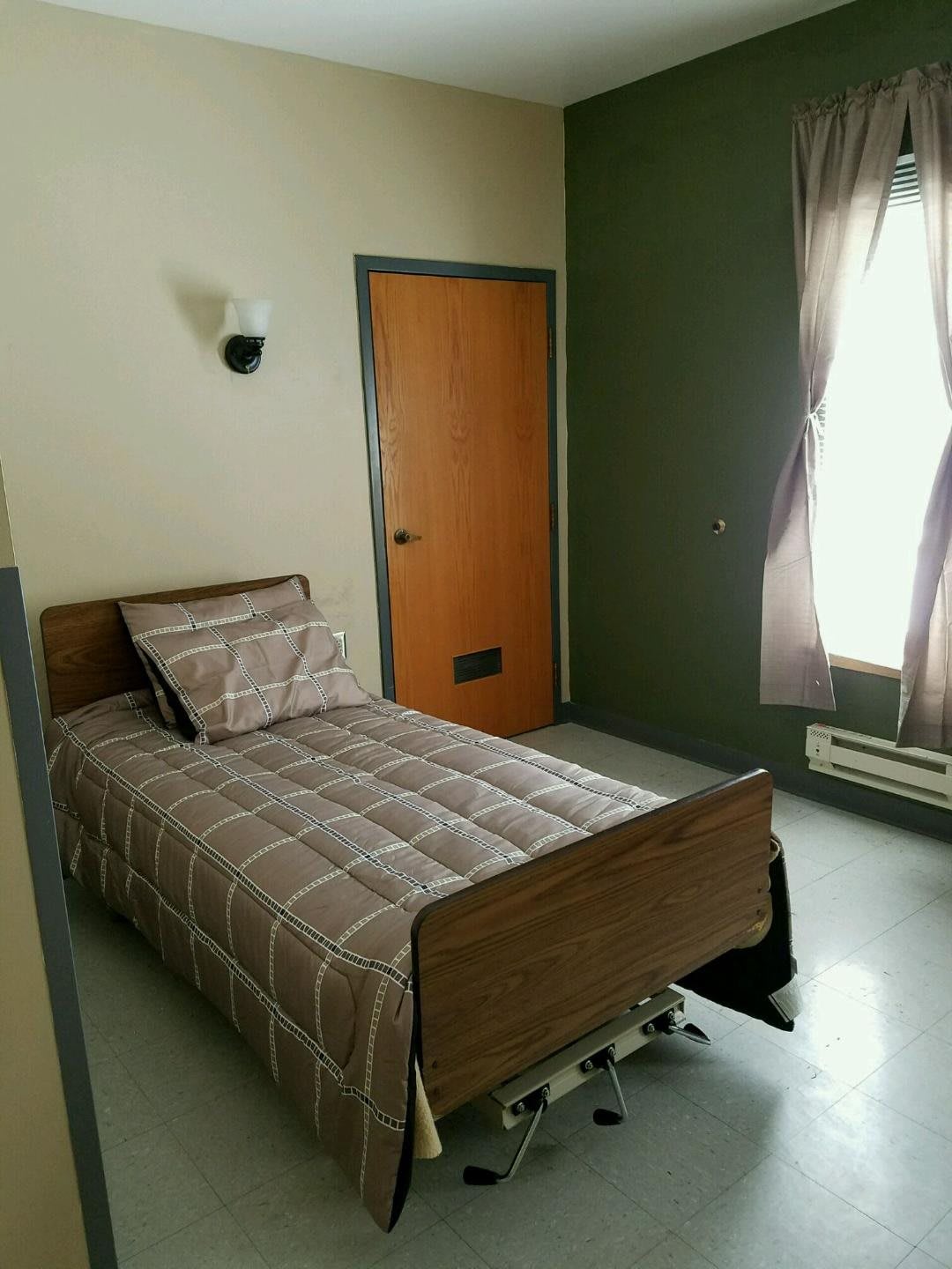 A minimalist bedroom featuring a single bed with plaid bedding, a window with curtains, and a wooden door.