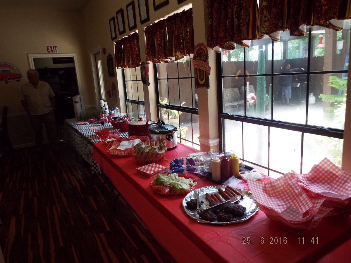 A buffet setup with various foods on a table covered with a red tablecloth next to large windows.