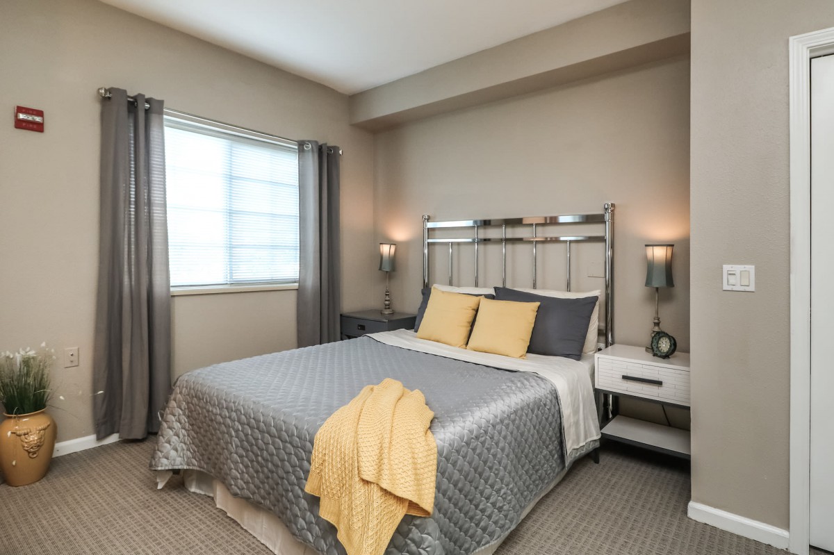 A modern bedroom with a neatly made bed featuring gray and yellow accents, complemented by bedside lamps and a window with curtains.