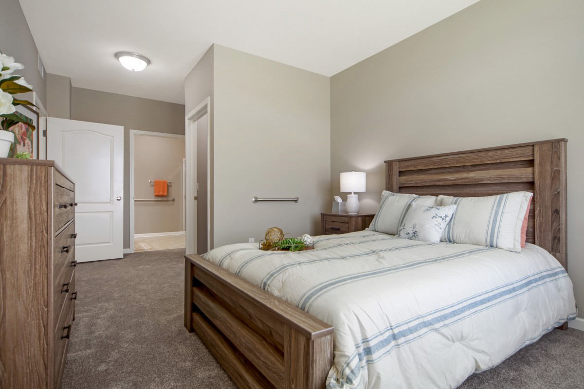A modern bedroom interior featuring a wooden bed, lamp, and soft carpeting.
