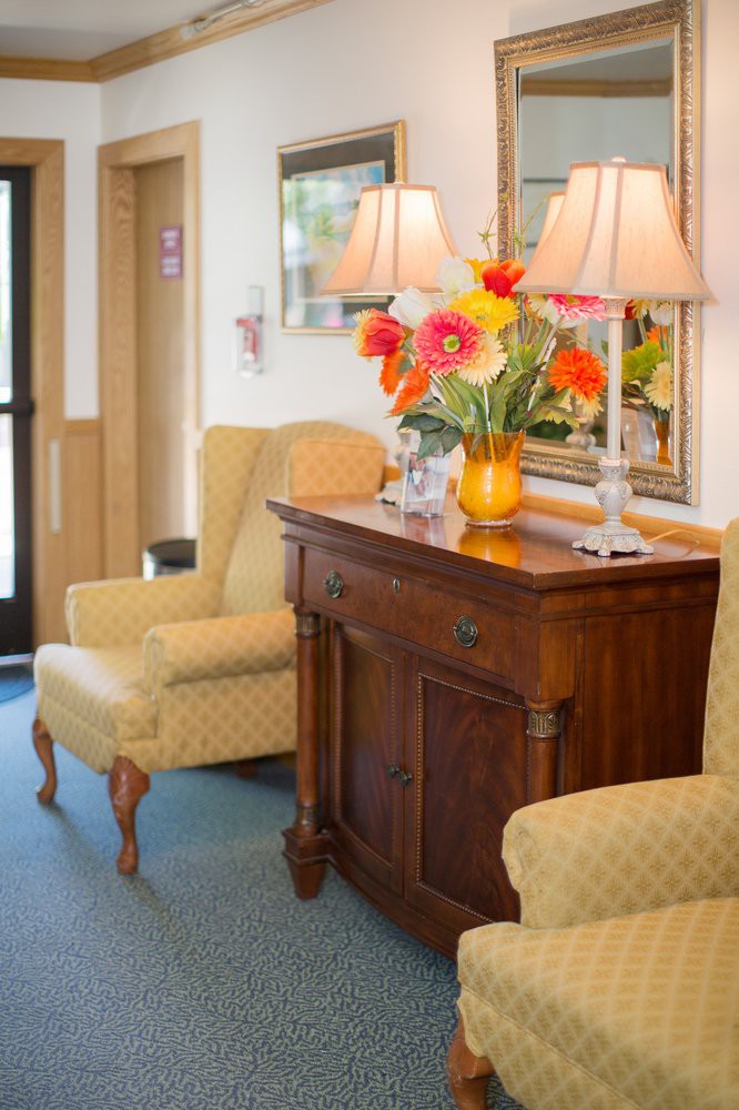 A cozy living room corner with upholstered chairs, a wooden cabinet topped with vibrant flowers and lamps.