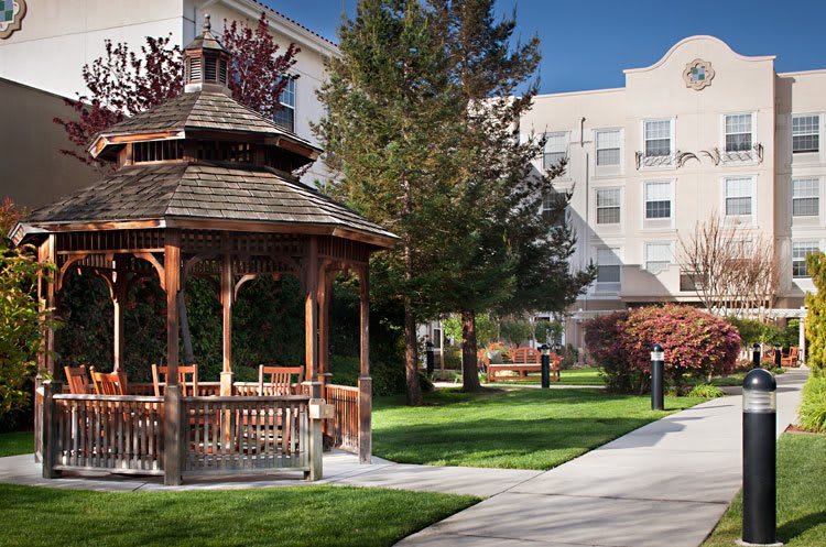A peaceful outdoor scene featuring a wooden gazebo surrounded by a lush garden and a walkway leading to a nearby building.