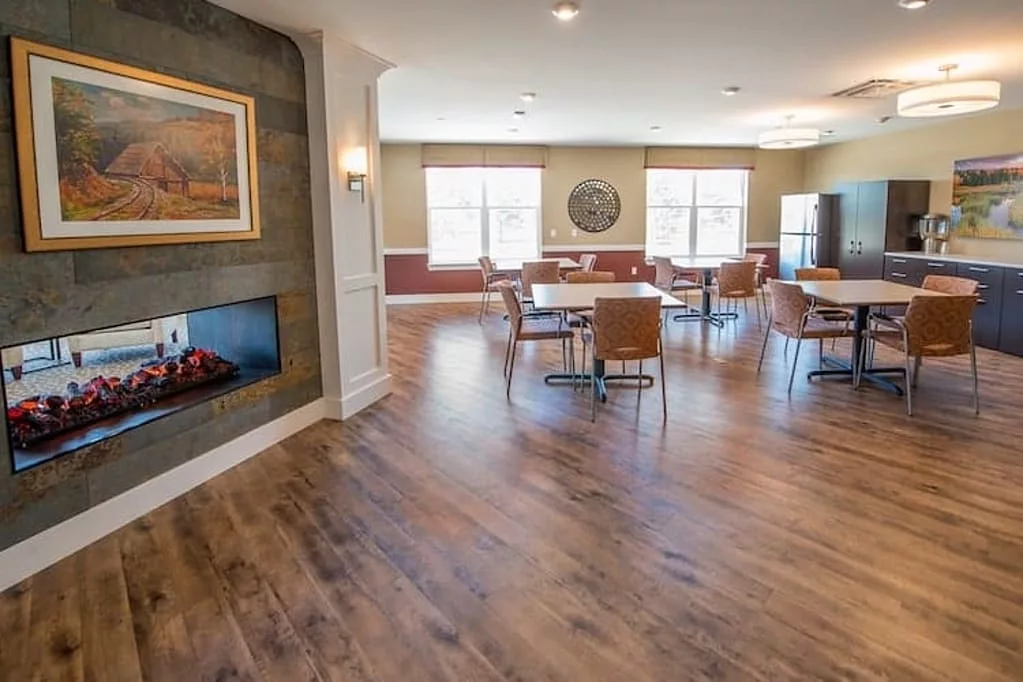 A cozy dining space with wooden floor, featuring a fireplace and ample natural light from large windows.