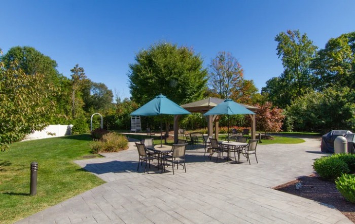 A sunny outdoor patio area with tables, chairs, and umbrellas surrounded by lush green landscaping.