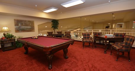 A luxurious billiard room featuring a pool table with red felt, elegant chairs, and plush red carpeting.