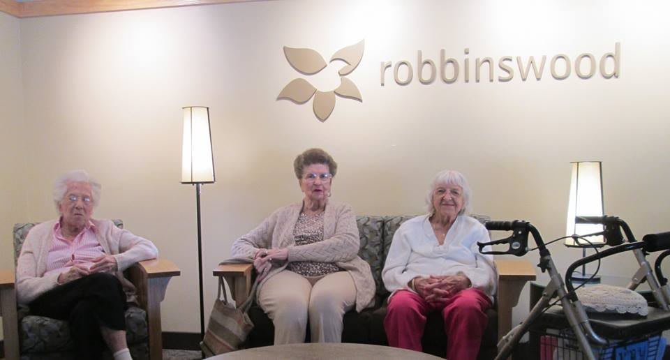 Two elderly individuals seated in a retirement home lounge area under a logo.