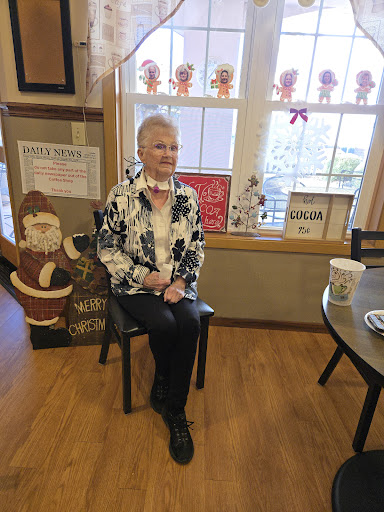 An elderly woman seated indoors, surrounded by a cozy, decorated atmosphere.