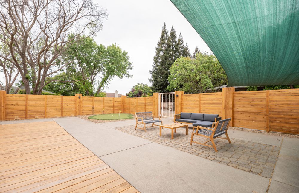 A spacious patio with modern outdoor furniture and a shade sail, surrounded by wooden fencing and trees.