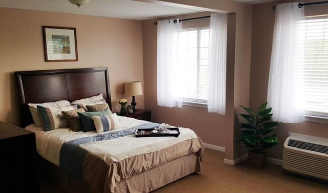 A cozy bedroom with a neatly made bed, bedside lamp, and a window letting in natural light.