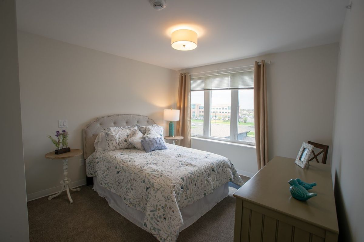A cozy bedroom with a neatly made bed, a lamp, and a window letting in natural light.