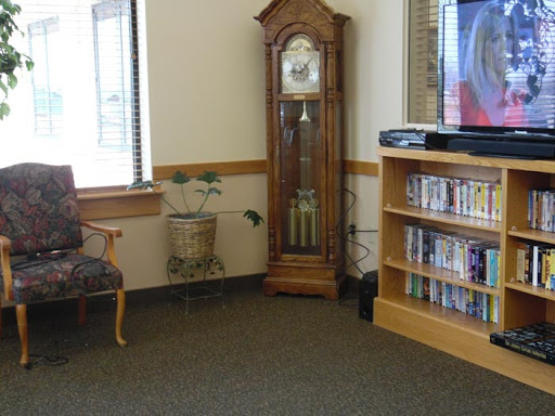 A cozy waiting room featuring an antique clock, a comfortable chair, a bookshelf filled with videos, and a television.