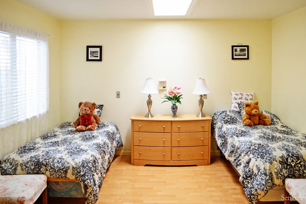 A cozy bedroom featuring twin beds with matching bedding, decorated with teddy bears and a wooden dresser centered between them, adorned with lamps and flowers.