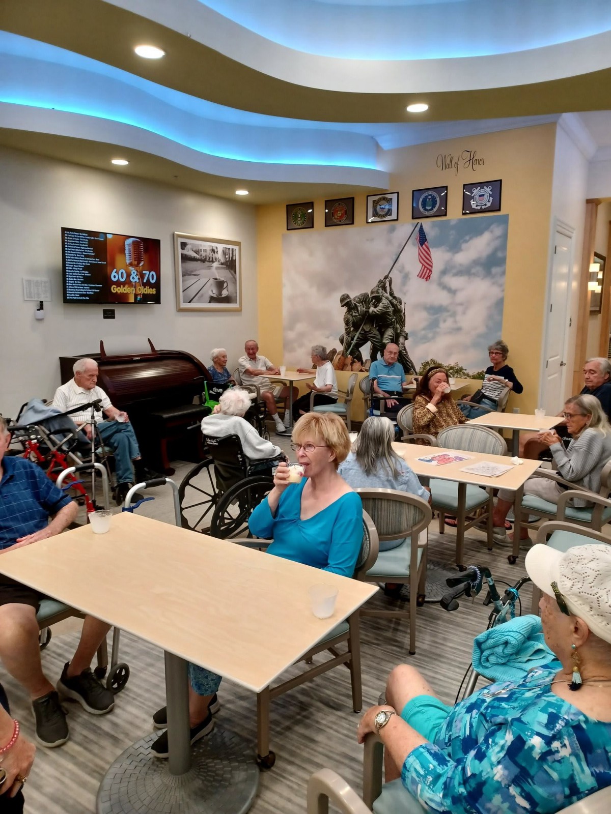 A community room filled with elderly individuals gathered around tables, engaging in a social activity. An American-themed mural decorates the background.