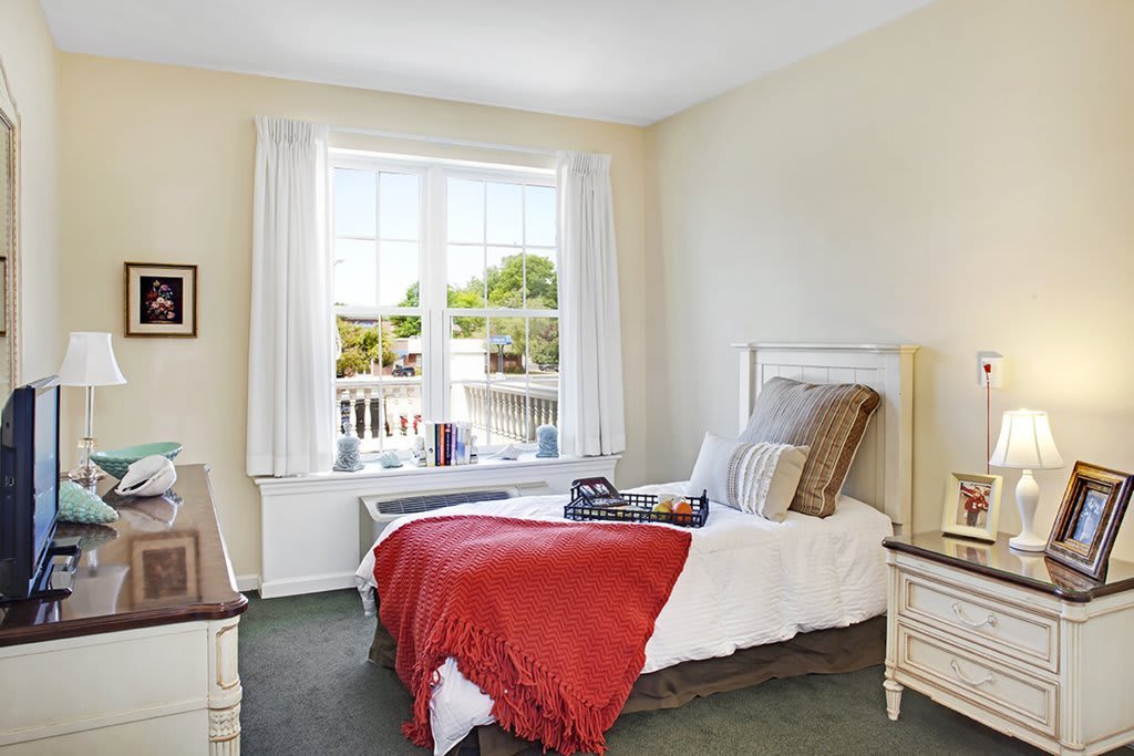 A cozy bedroom with a single bed, accented with a red blanket, and natural light streaming in through the window.