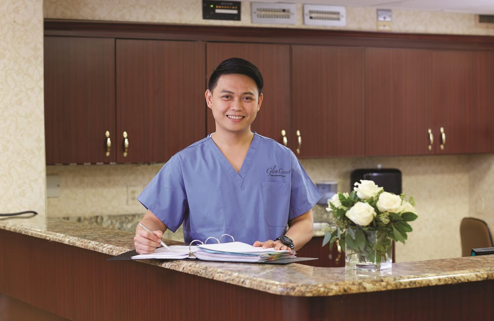 A medical professional in blue scrubs stands at a reception desk with a clipboard.