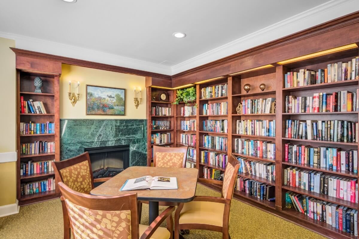 A cozy library room with bookshelves, a table, chairs, and a fireplace.