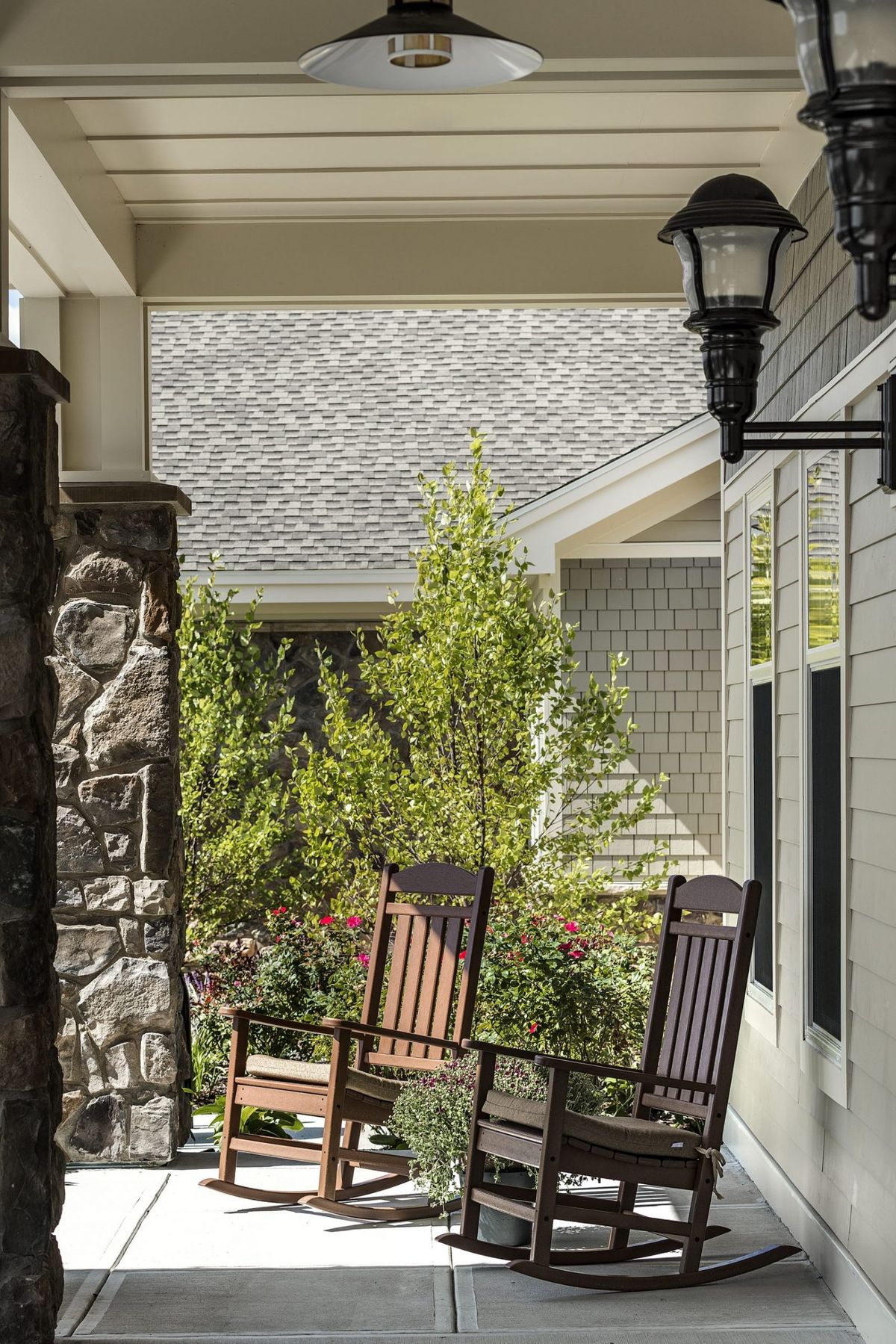 A cozy porch with wooden rocking chairs next to a stone pillar, adorned with lanterns and surrounded by lush greenery.