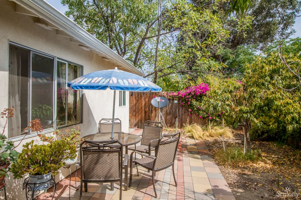 A cozy outdoor patio area with a table, chairs, and an umbrella, surrounded by greenery and flowers.