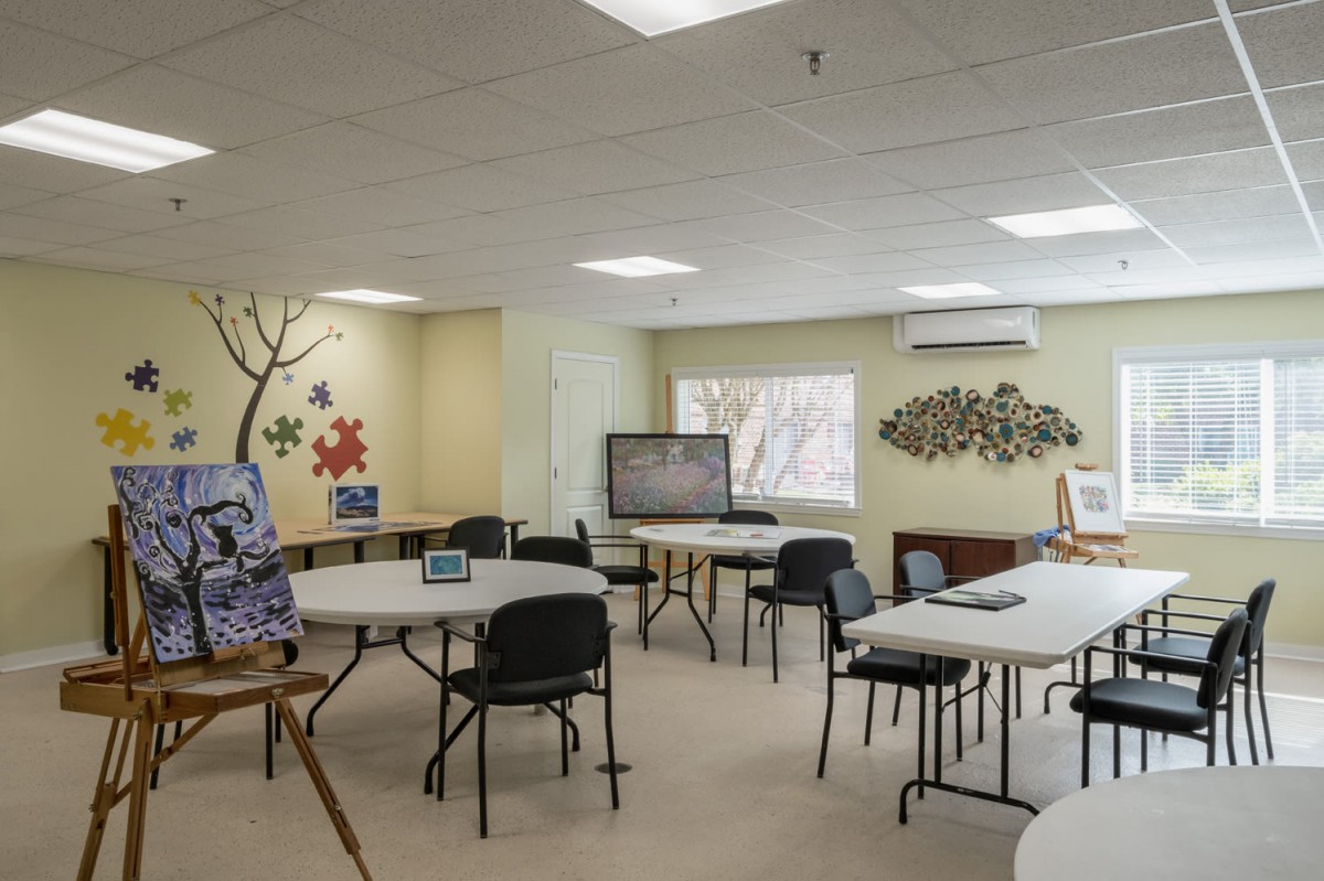 A community meeting room with tables and chairs arranged for group activities, featuring wall decorations and natural light from a window.