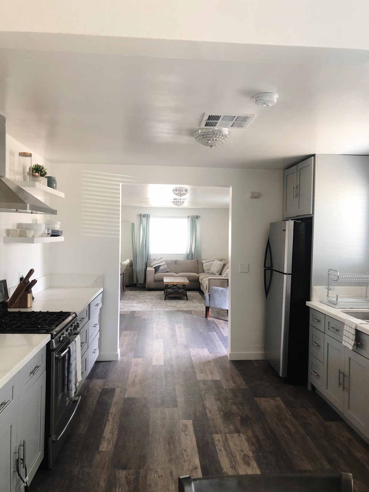 A modern kitchen with hardwood floors, white cabinetry, and stainless steel appliances leading into a bright living room.