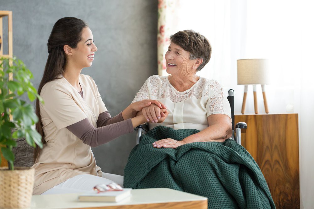 A caregiver smiling and holding the hand of an elderly woman in a wheelchair.