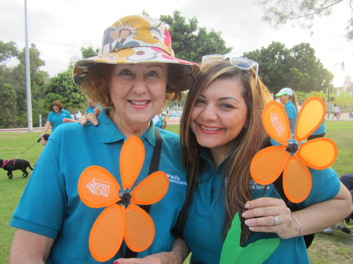 Two women smiling and holding orange flower decorations at an outdoor event.