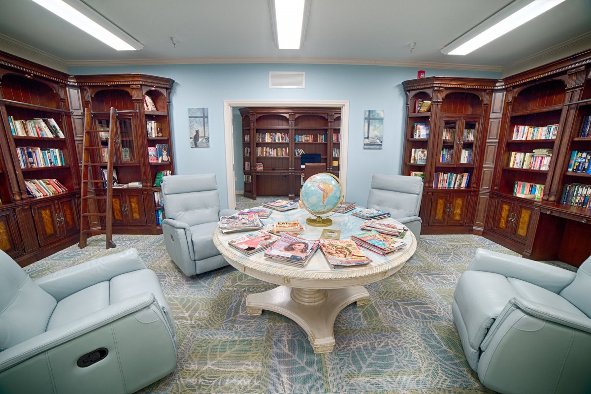 A cozy library corner with reclining chairs, a central table displaying magazines, and wooden bookshelves lining the walls.
