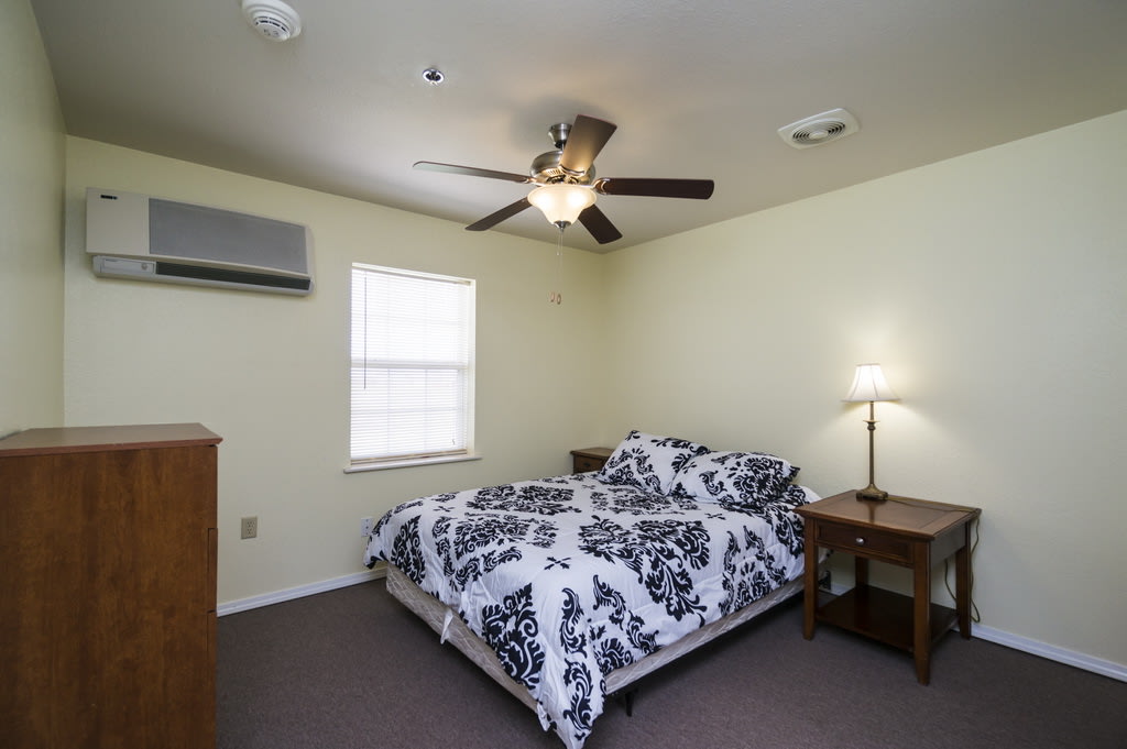 A cozy bedroom with a patterned bedspread, ceiling fan, and a nightstand lamp.