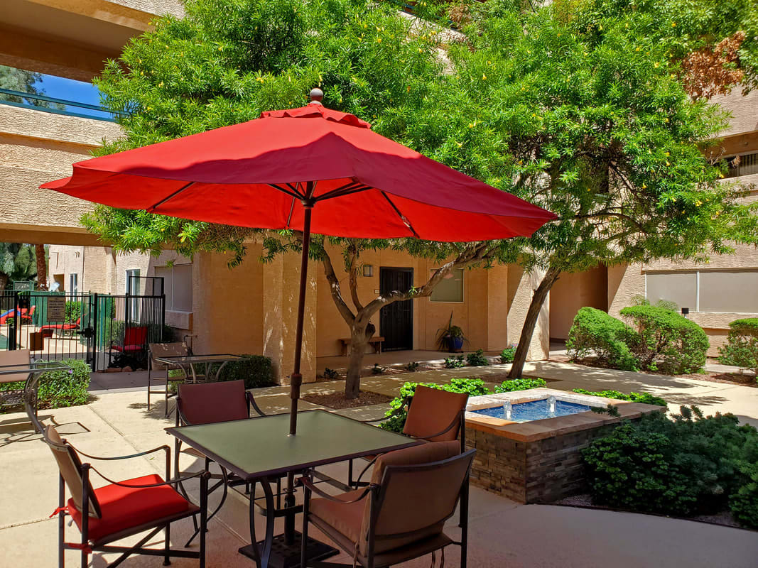 A sunny courtyard with a red umbrella shading a table and chairs, surrounded by trees and a small fountain.