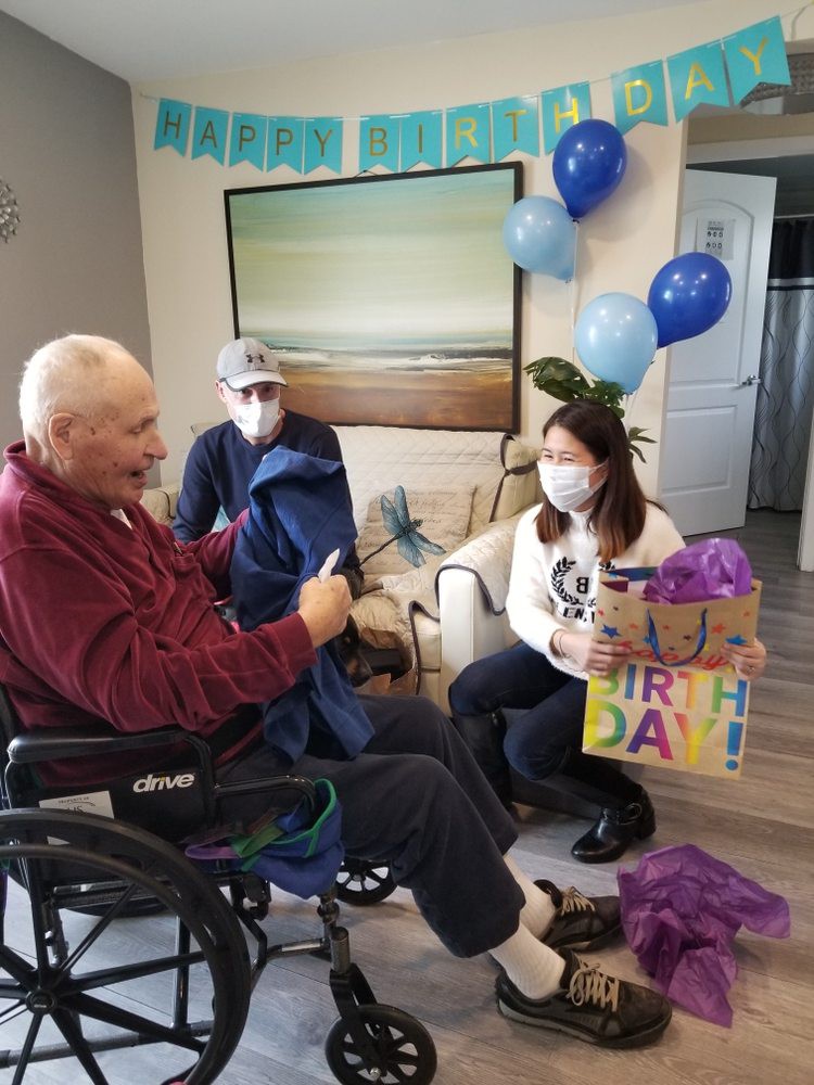An elderly man receives a birthday gift from family members during a celebration at home.