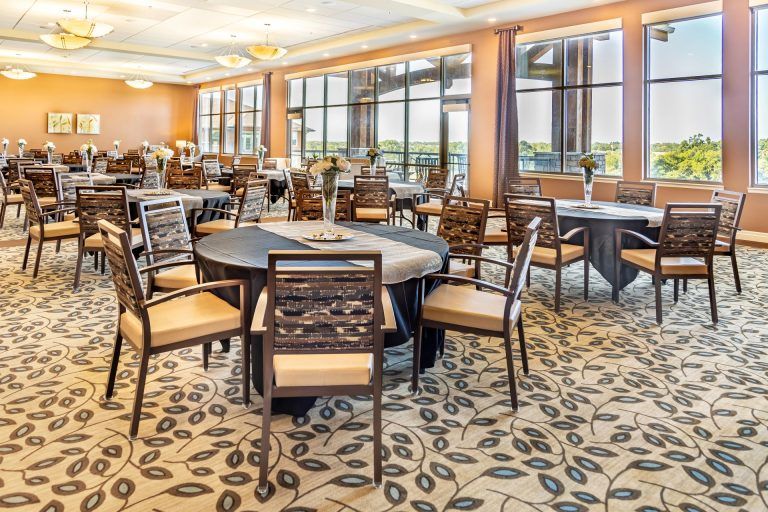 A spacious dining area with round tables and cushioned chairs, featuring large windows and a patterned carpet.