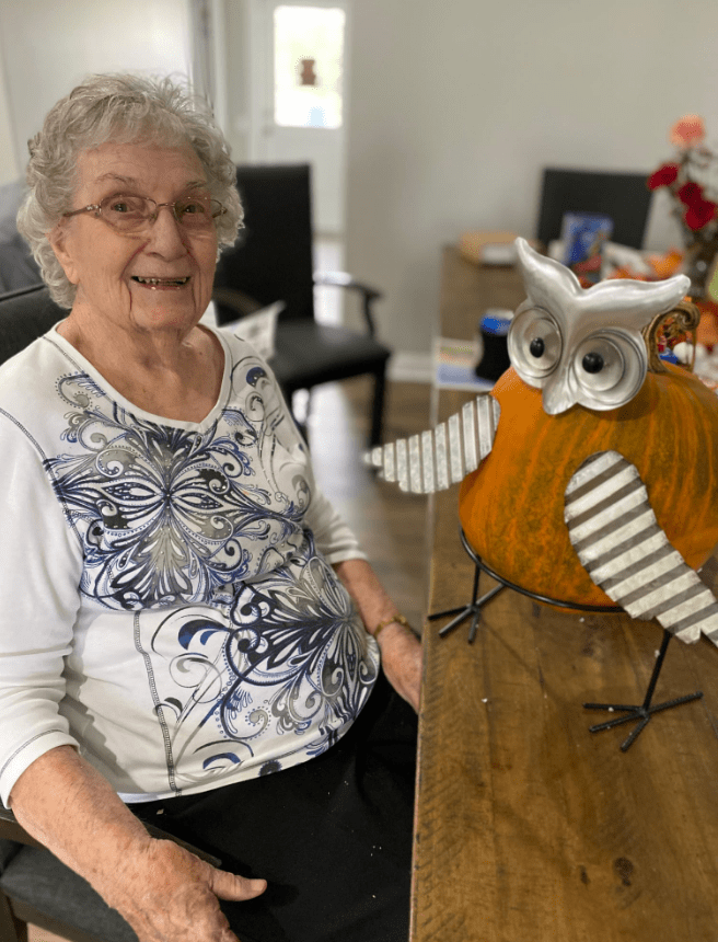 An elderly woman sitting indoors next to a decorative owl figurine, smiling at the camera.