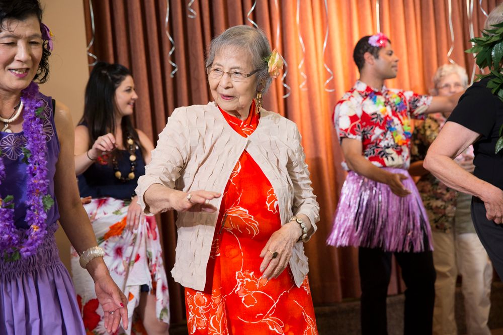 An elderly woman joyfully dances at a tropical-themed party, surrounded by others in colorful outfits.
