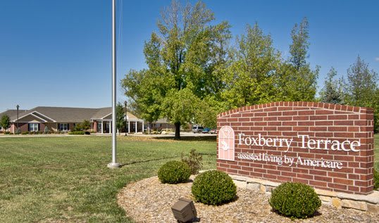 A landscaped entrance with a sign for Foxberry Terrace Assisted Living by Americare, featuring a well-maintained lawn and trees on a clear day.