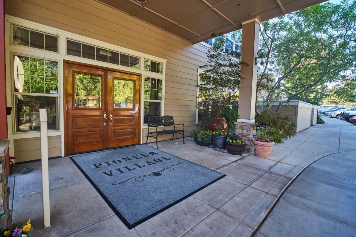 Entrance of a building with wooden doors, a mat labeled 'Pioneer Village,' and potted plants.
