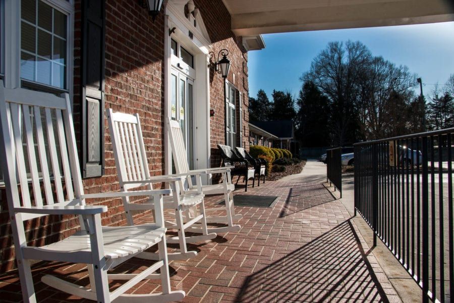 A sunny porch features white rocking chairs against a brick house.