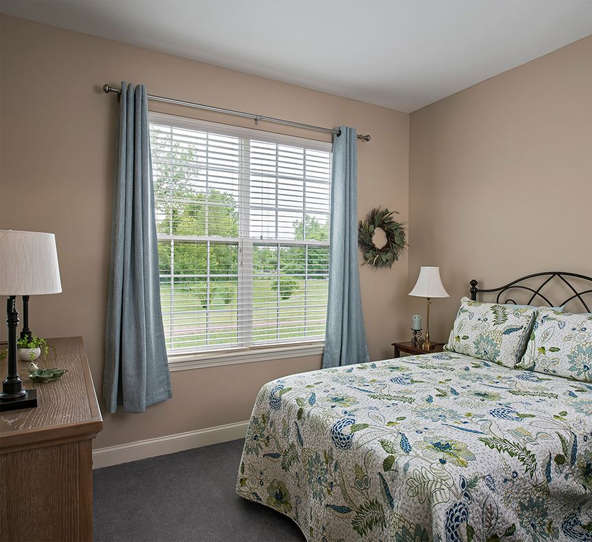 A cozy bedroom with floral bedding, a window with blue curtains, and soft lighting.
