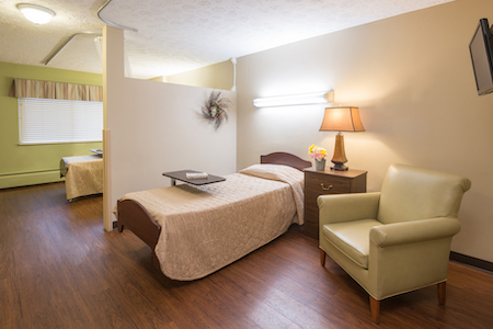 A cozy and well-lit bedroom featuring a neatly made bed, a bedside table with a lamp, and a comfortable chair.