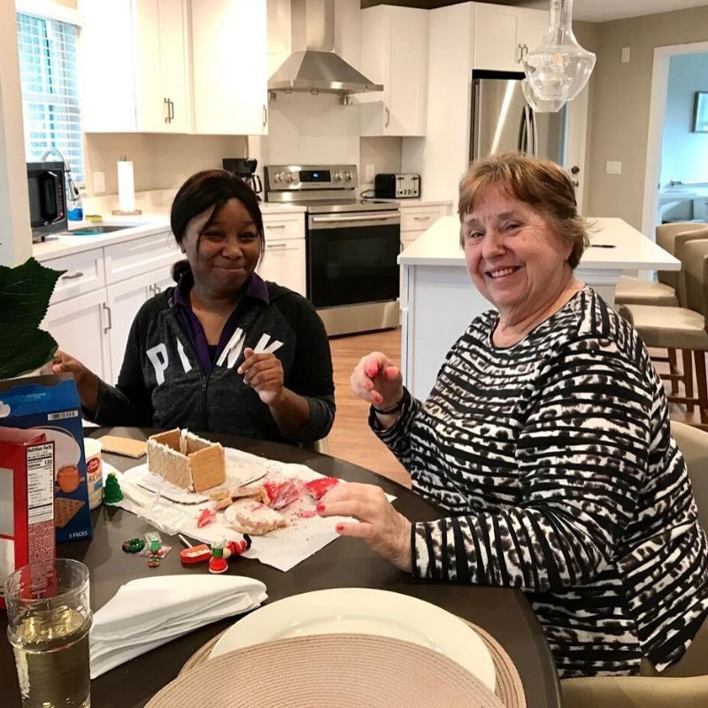 Two women smiling while making a gingerbread house in the kitchen.