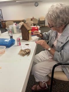 An elderly woman is engaged in painting a gingerbread house while seated at a table.
