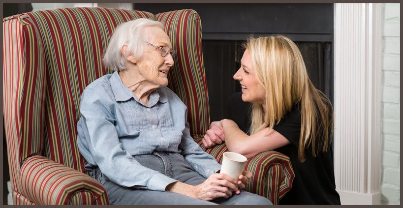 An elderly woman and a younger woman smiling and having a conversation in a cozy setting.