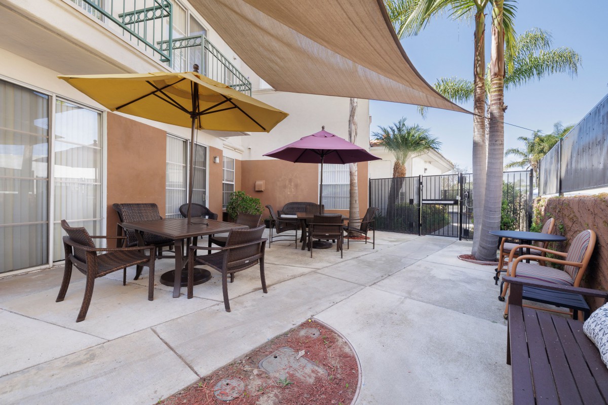 A cozy outdoor patio area with tables and chairs under colorful umbrellas and shade sails, surrounded by palm trees.