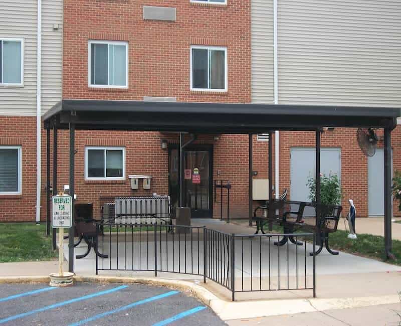 The image shows the entrance of a building with a covered area featuring benches, surrounded by a small fence, and adjacent to a marked parking space.