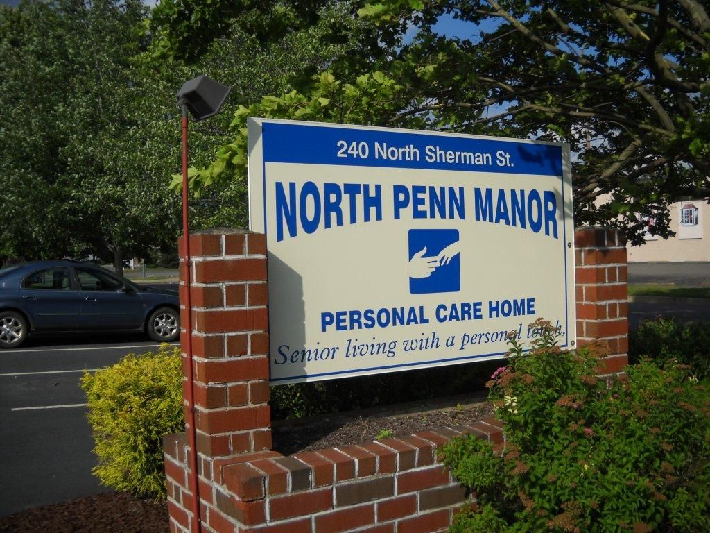 Sign for North Penn Manor Personal Care Home on a brick wall surrounded by greenery.