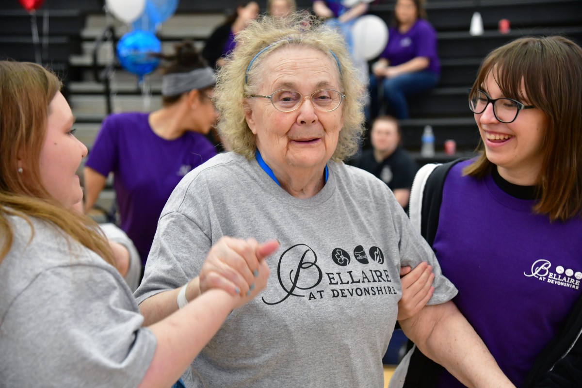 An elderly woman participates in a community sports event, holding hands with others and smiling warmly.