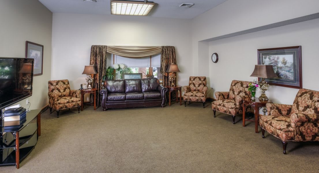 A cozy living room with a sofa, armchairs, and a large window adorned with curtains and indoor plants.