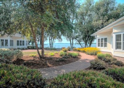 A serene garden pathway leads to a picturesque lakeside view, surrounded by lush greenery and charming buildings.